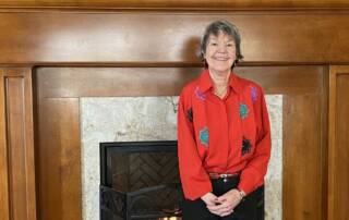 Smiling senior woman in a red shirt standing by a fireplace.